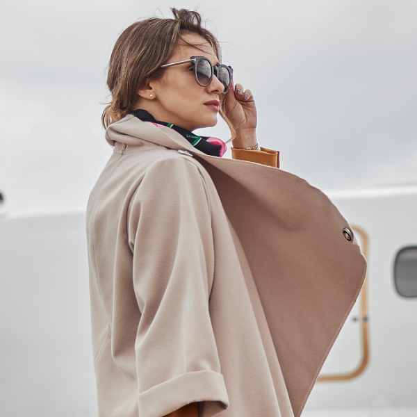 A person in a beige coat and sunglasses stands outside near an airplane on a cloudy day, pondering the possibilities of aircraft acquisition.