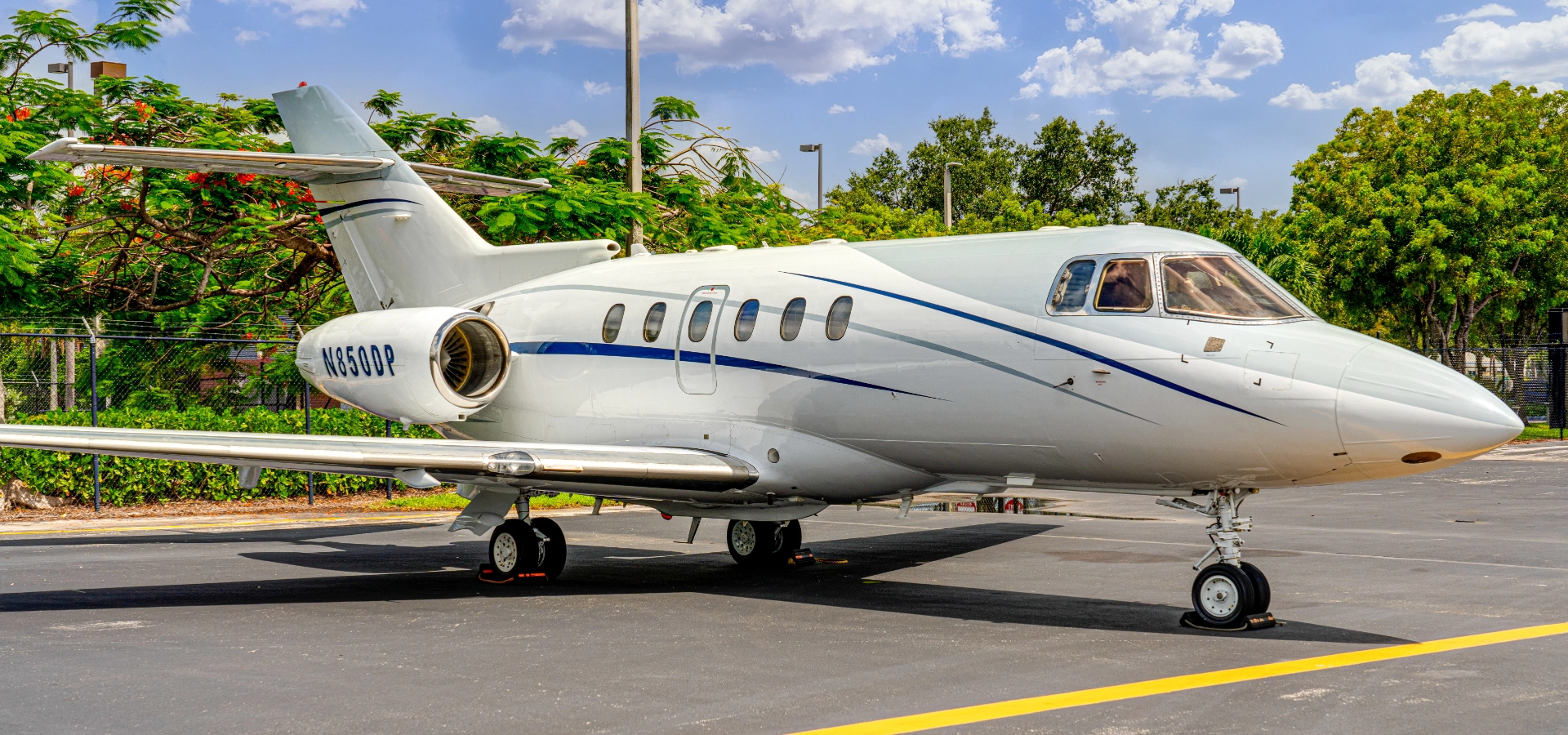A luxurious private jet, part of a prime collection in the business jets for sale market, sits boldly on an airport tarmac, framed by lush trees and a partly cloudy sky.