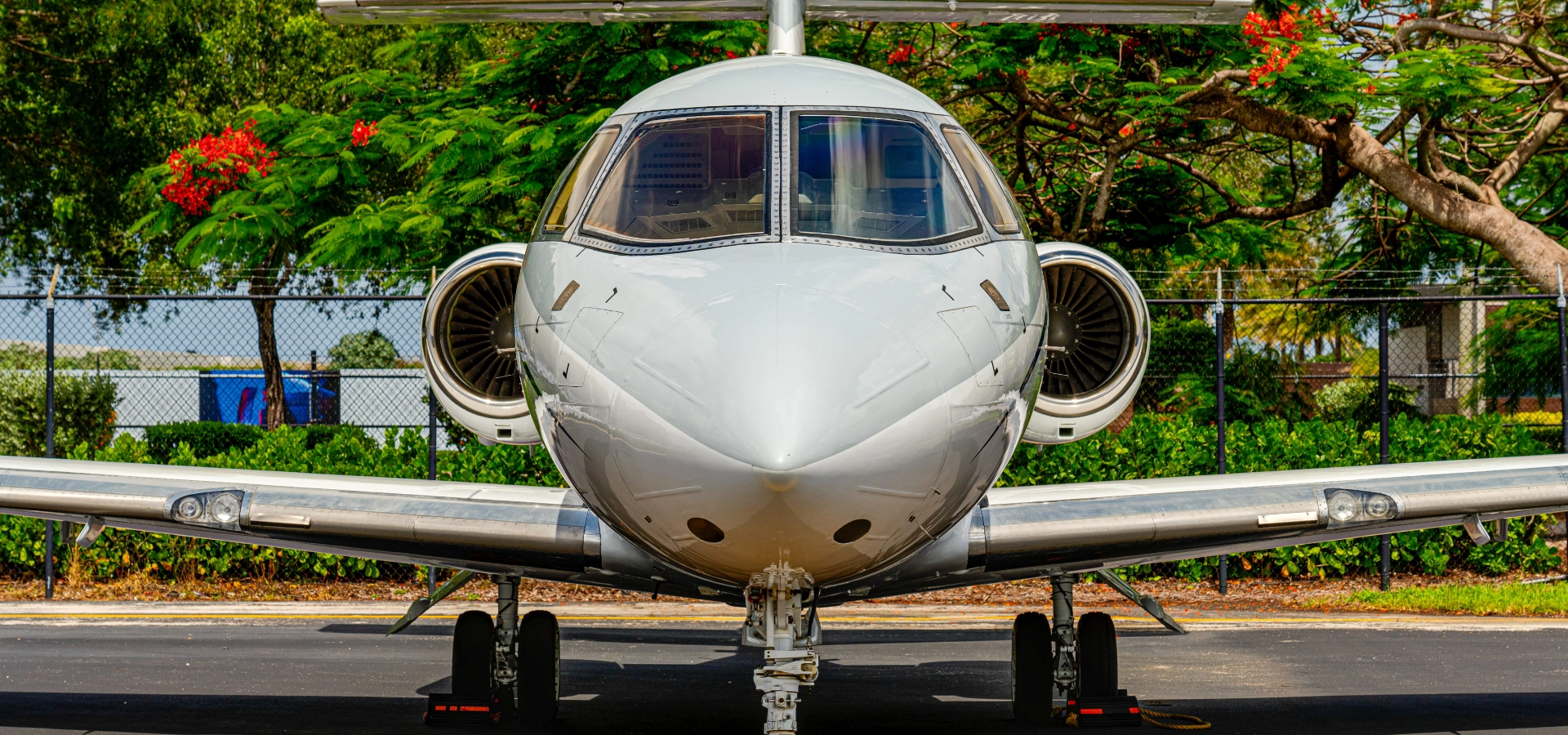 Front view of a parked white private jet with engines visible on both sides, exuding elegance amidst the greenery and fence in the background—a true testament to luxury jet sales.
