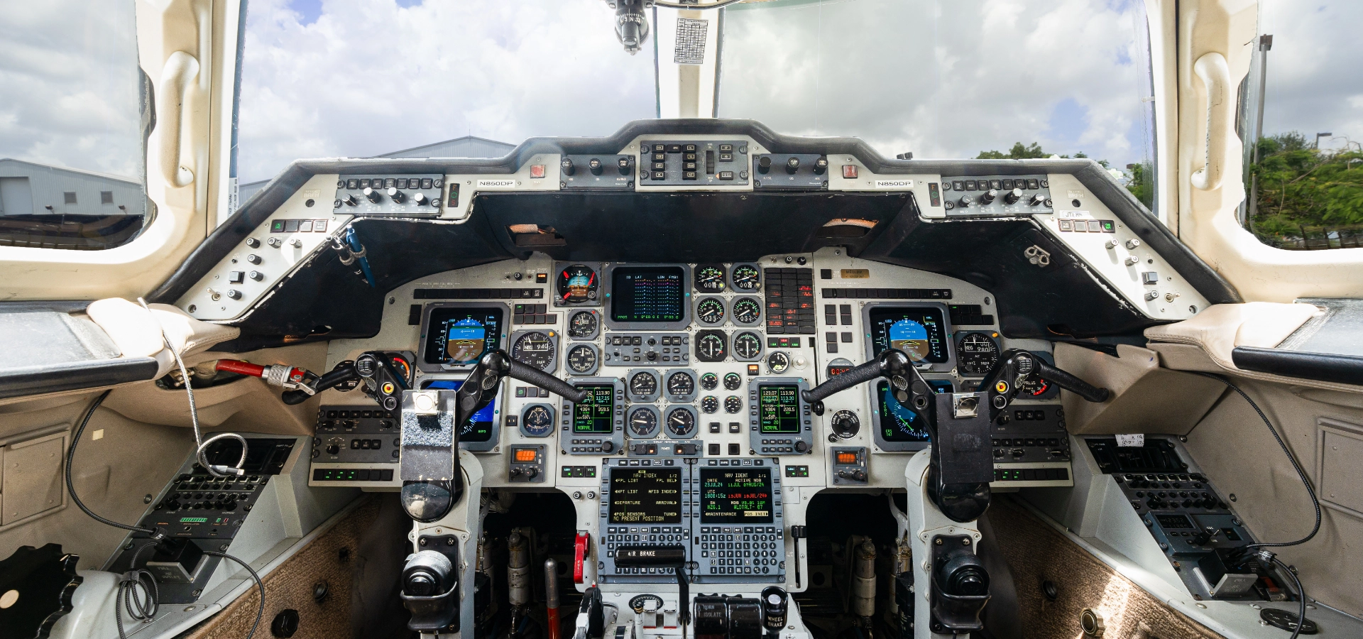 Interior view of an aircraft cockpit featuring various control panels, instruments, and two pilot seats. The scene captures the essence of luxury jet sales with a cloudy sky visible through the windshield, inviting you to explore high-end aircraft acquisition.