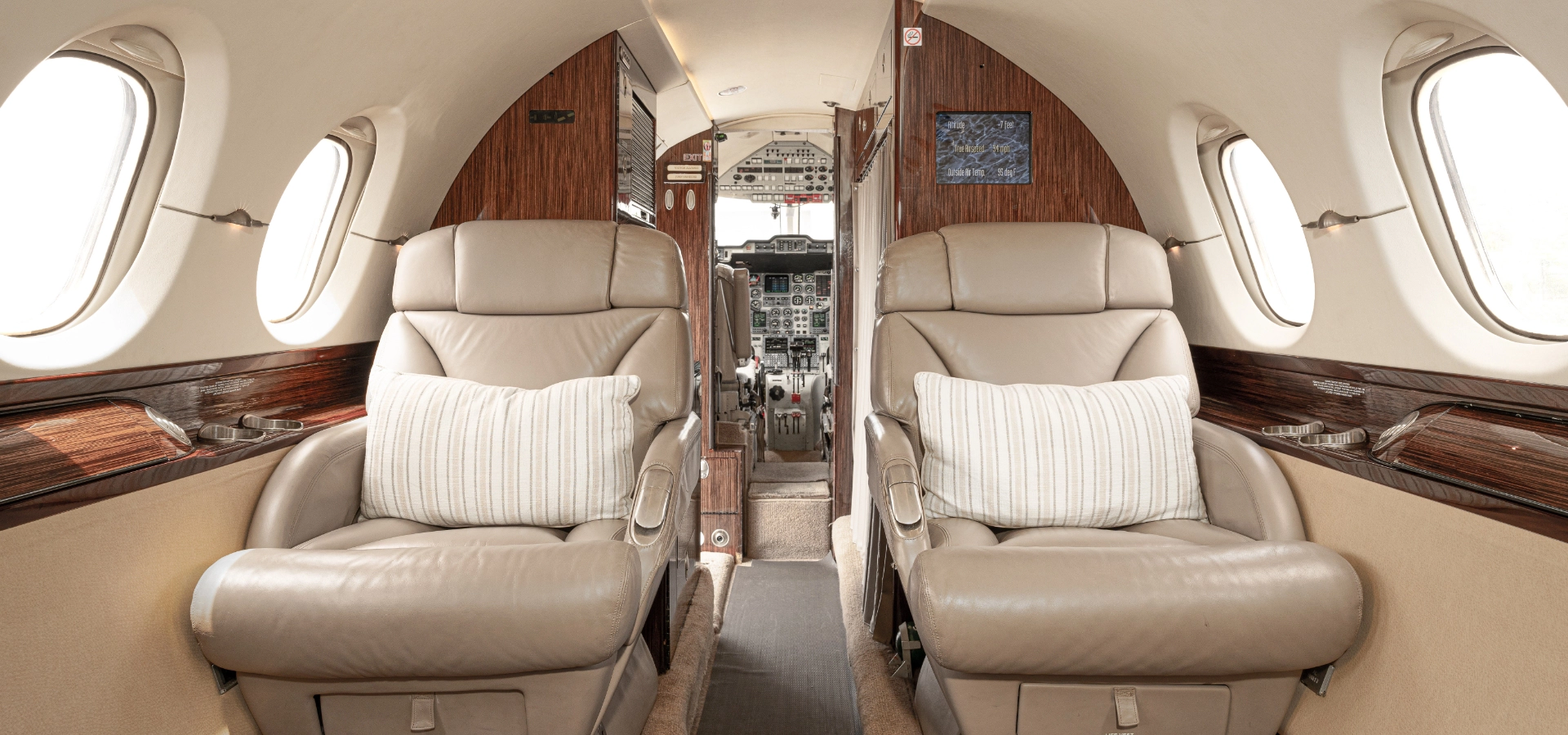 Explore the epitome of opulence with this impeccable private jet interior, featuring two beige leather seats, elegant wood paneling, and a captivating view of the cockpit. Perfect for discerning buyers in luxury jet sales seeking unparalleled comfort and style.