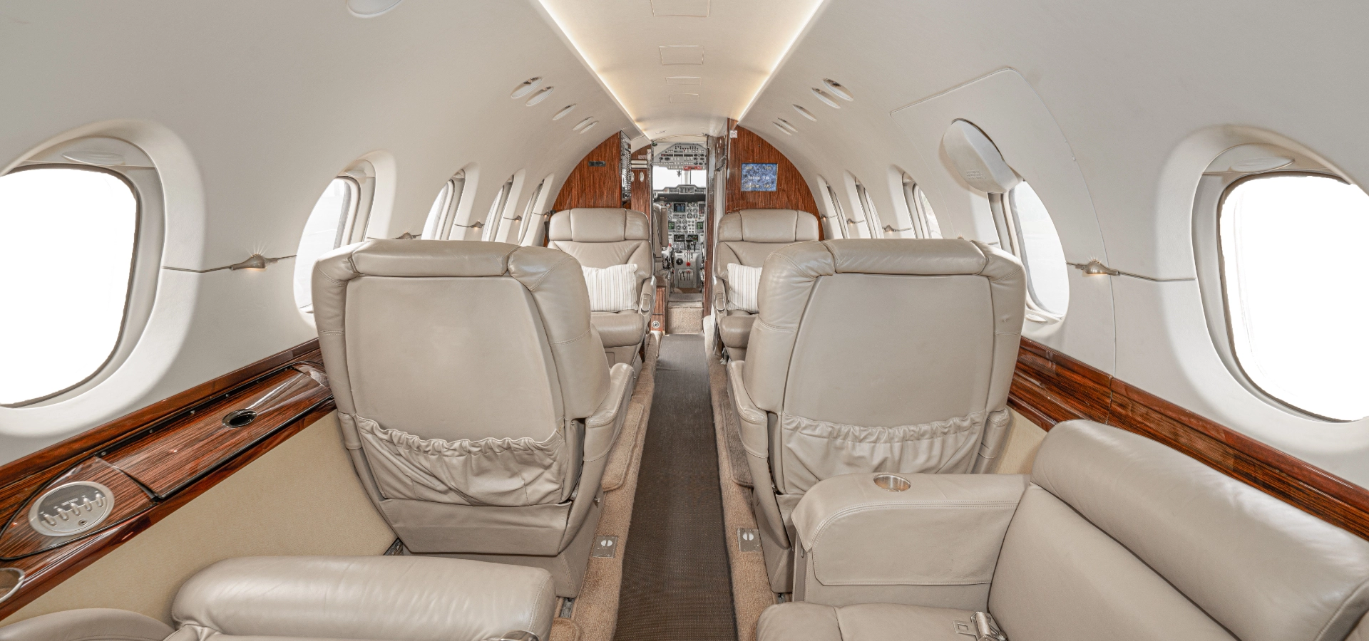 Explore the luxurious interior of a private jet with elegant beige leather seats and refined wood paneling, offering a seamless view towards the cockpit—exemplifying the excellence found in business jets for sale.