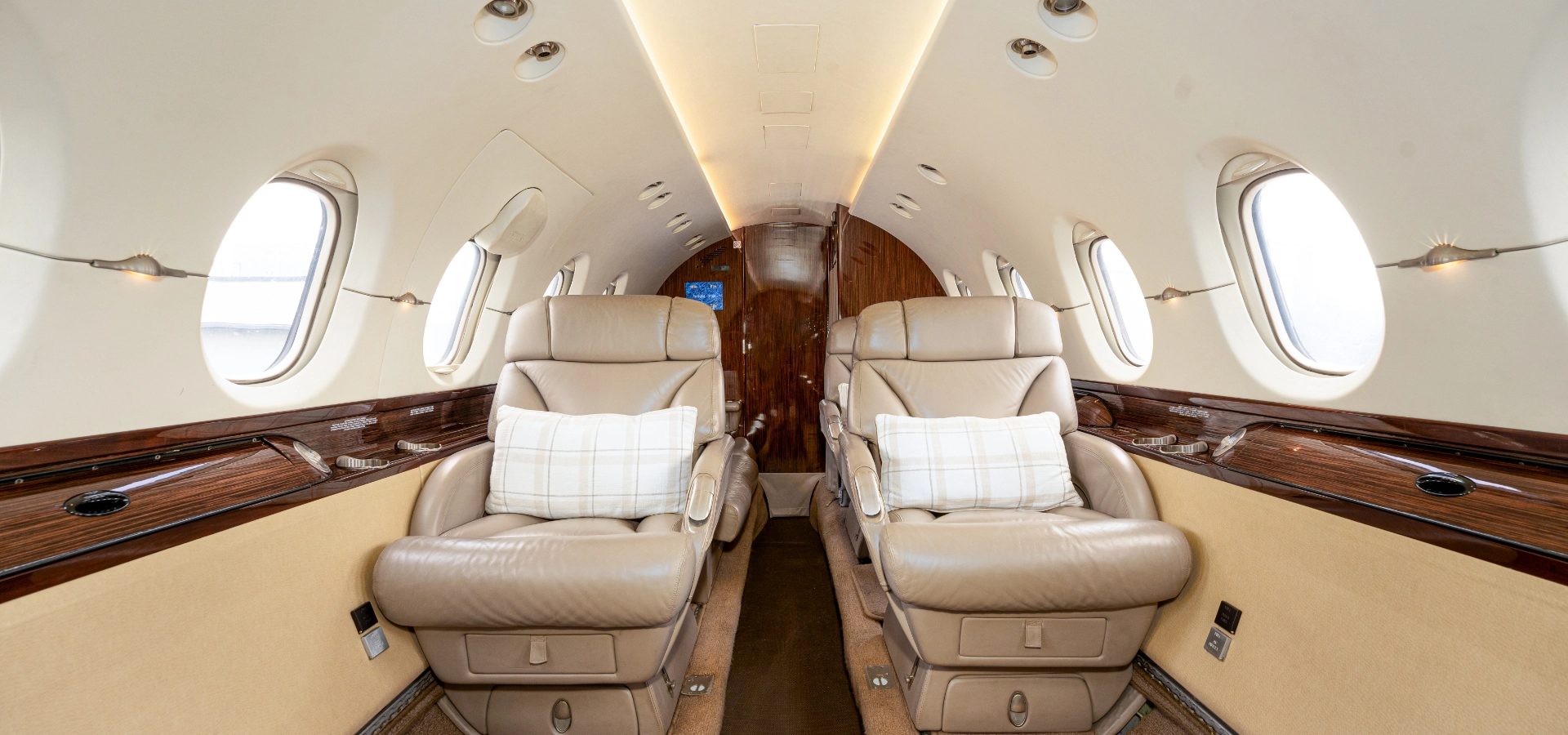 The interior of a private jet, perfect for those exploring aircraft acquisition, features two beige leather seats with plaid pillows, surrounded by wooden accents and oval windows.