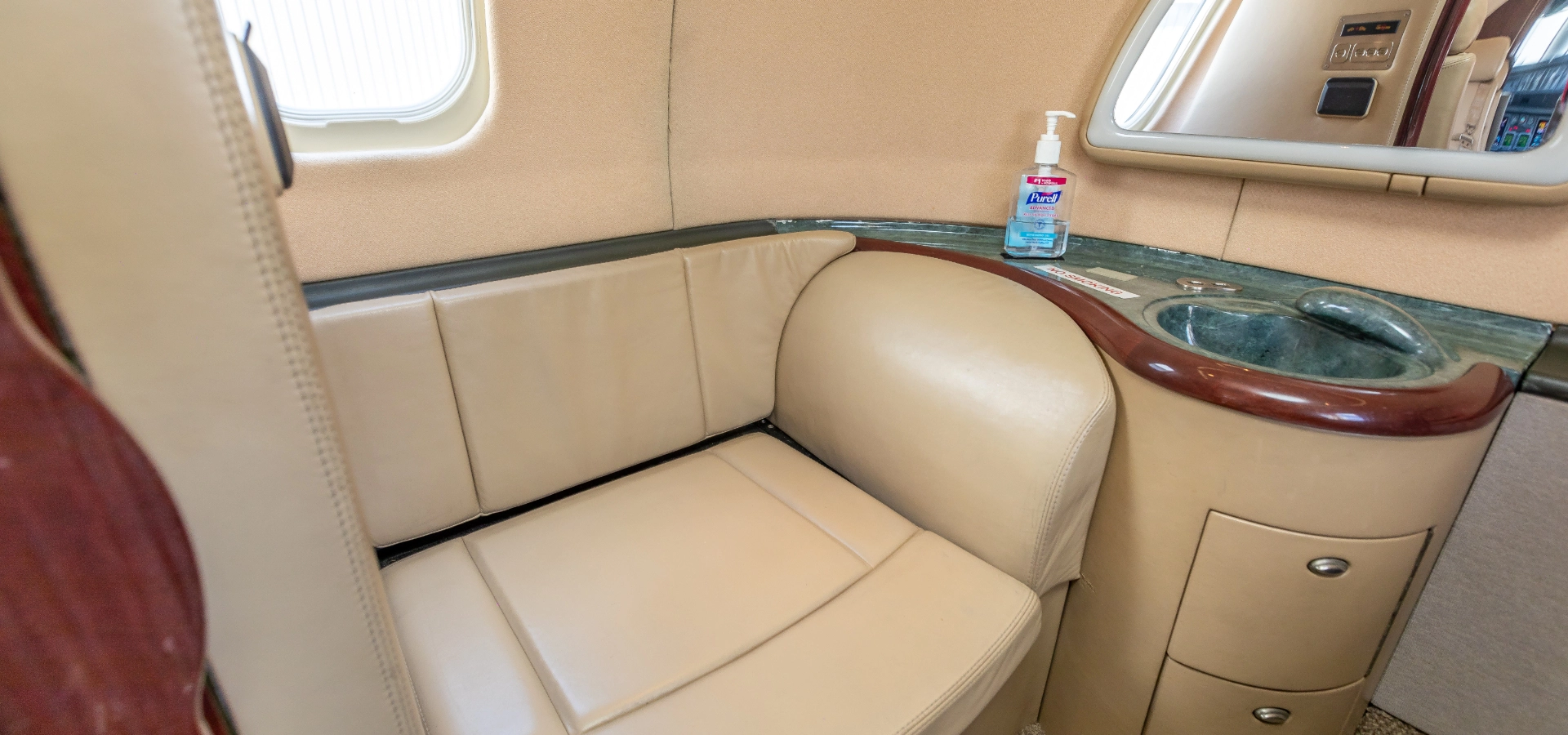 Close-up of a beige leather seat beside a wooden countertop with a built-in sink in an airplane cabin, highlighting the elegance found in luxury jet sales. A bottle of hand sanitizer sits neatly on the counter, exemplifying the refined comfort available through private jets for sale.