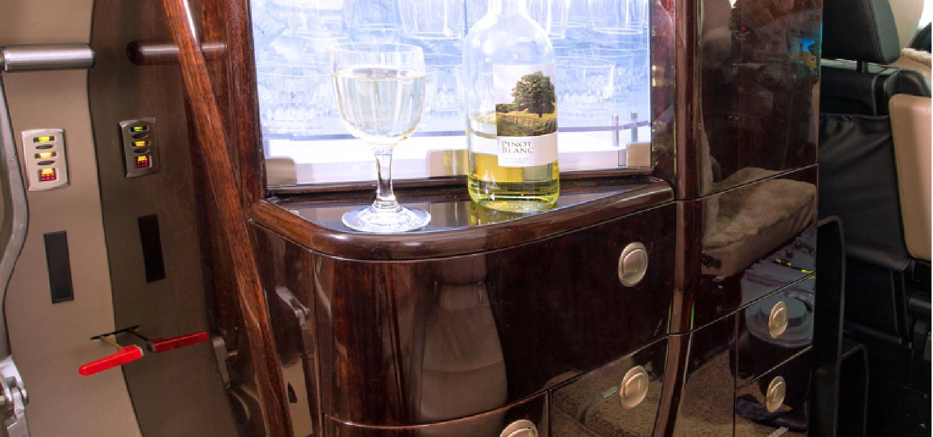 A bottle of white wine and a filled glass rest on a wooden surface with drawers, beside a window showcasing glassware, evoking the elegance synonymous with corporate jet sales.