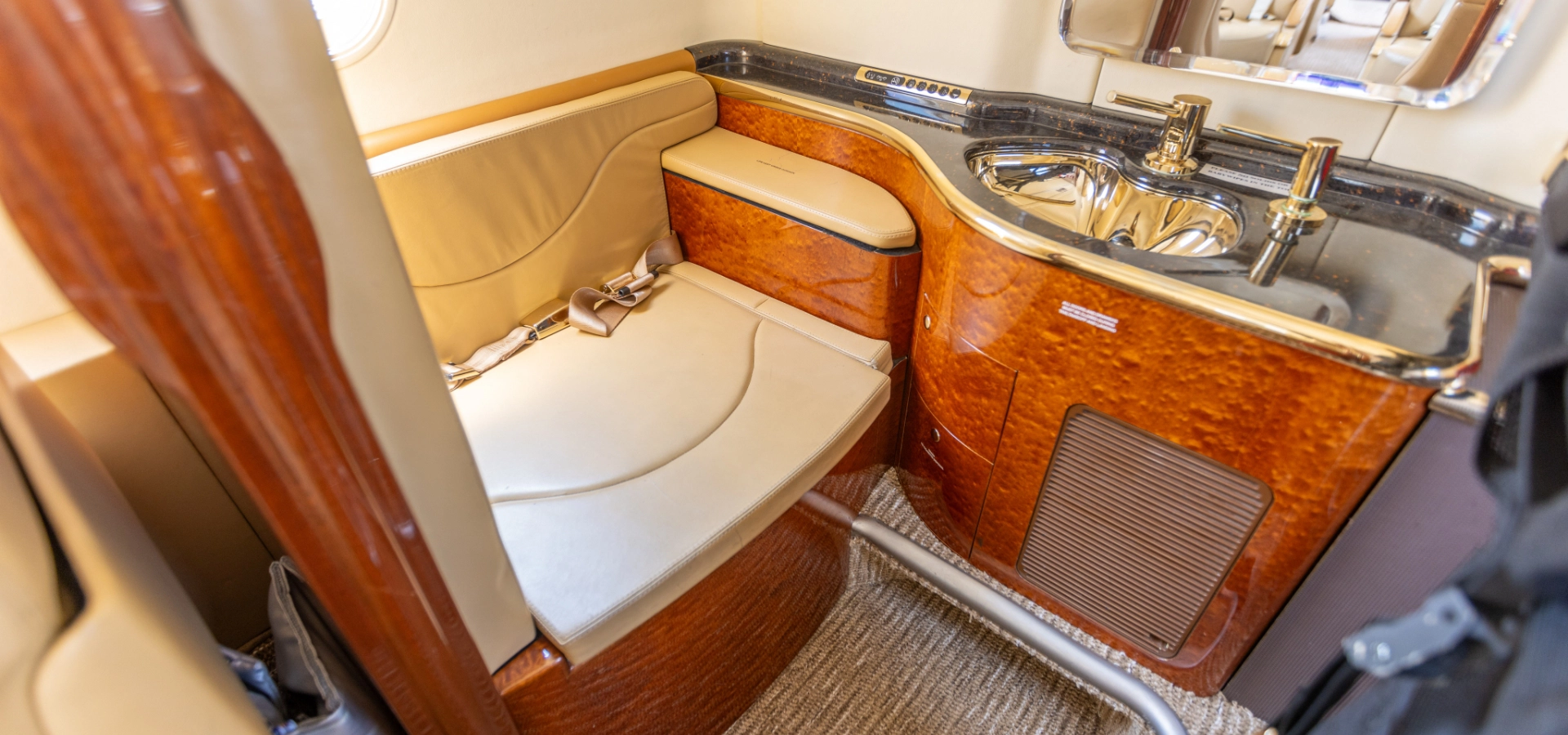 Luxurious private jet bathroom with wooden accents, a tan leather bench, gold faucet, and a mirrored cabinet—ideal for discerning buyers exploring business jets for sale.
