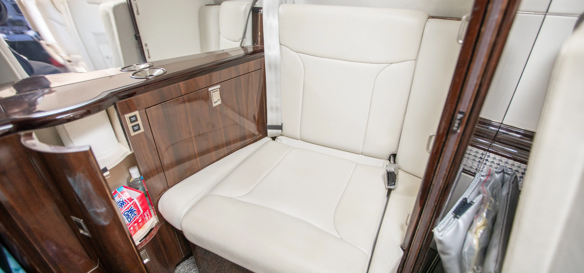 Luxurious airplane seat with white leather upholstery and a wooden panel side, exemplifying the elegance found in corporate jet sales. The visible seatbelt and a storage compartment packed with snacks and magazines complete this exquisite travel experience.