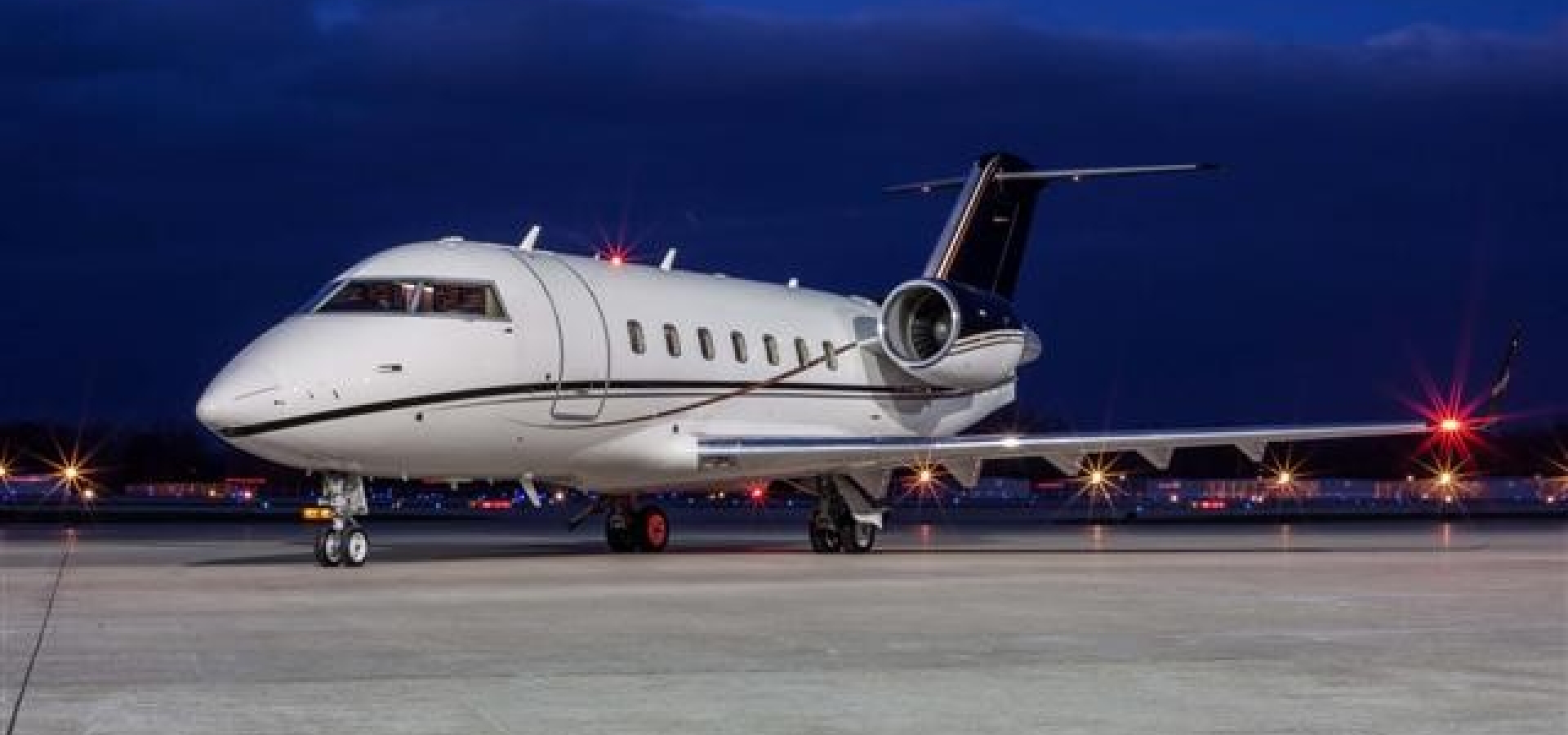 A private jet is parked on a dimly lit airport tarmac at night, with lights glowing in the background.
