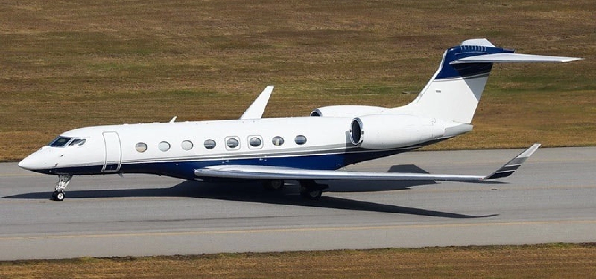 A sleek white and blue private jet, available through our corporate jet sales, gracefully taxis on a runway flanked by lush grassy areas.