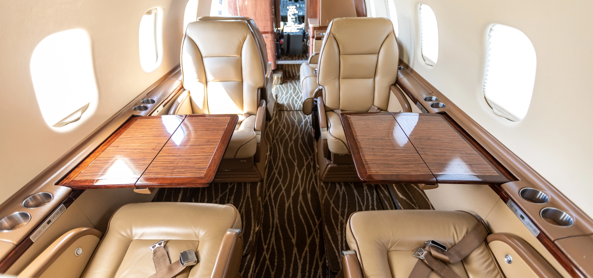 Explore the luxury of a private jet interior, with leather seats, fold-out wooden tables, and cup holders. The patterned carpet aisle leads gracefully to the cockpit, showcasing what you could own with our exclusive corporate jet sales options.