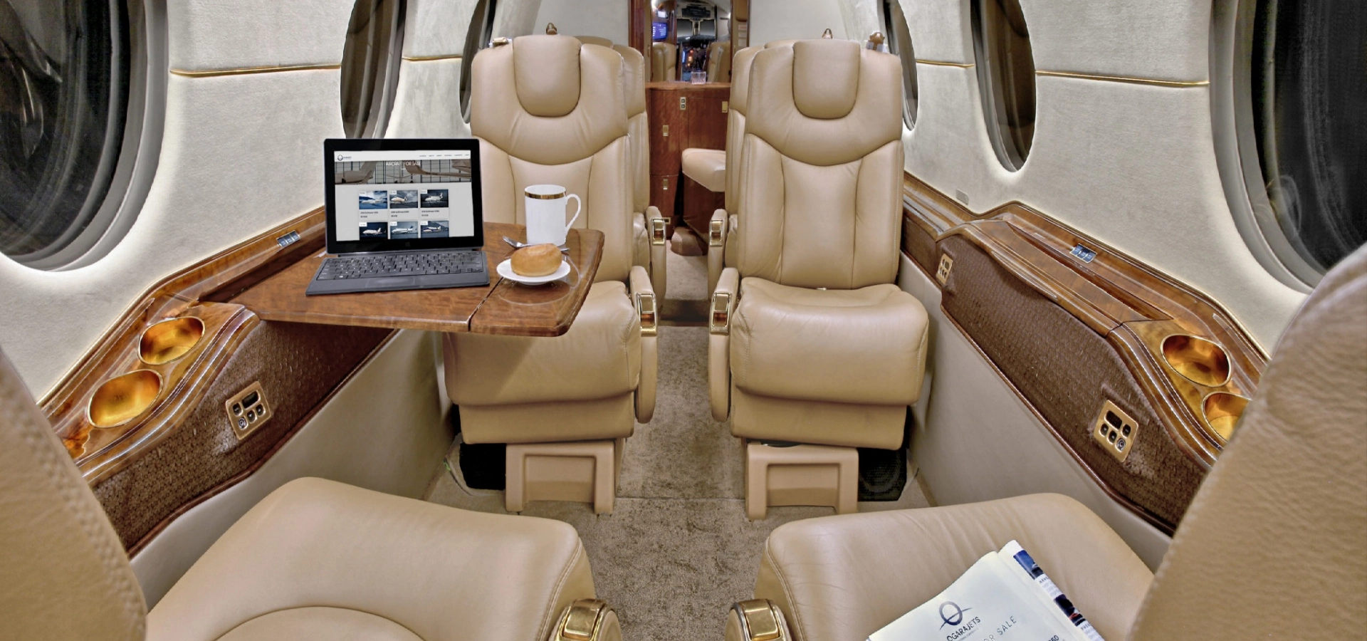 The interior of a private jet showcases beige leather seats, with a tray table elegantly holding a laptop and a cup of coffee—a perfect setting for those exploring private jets for sale.