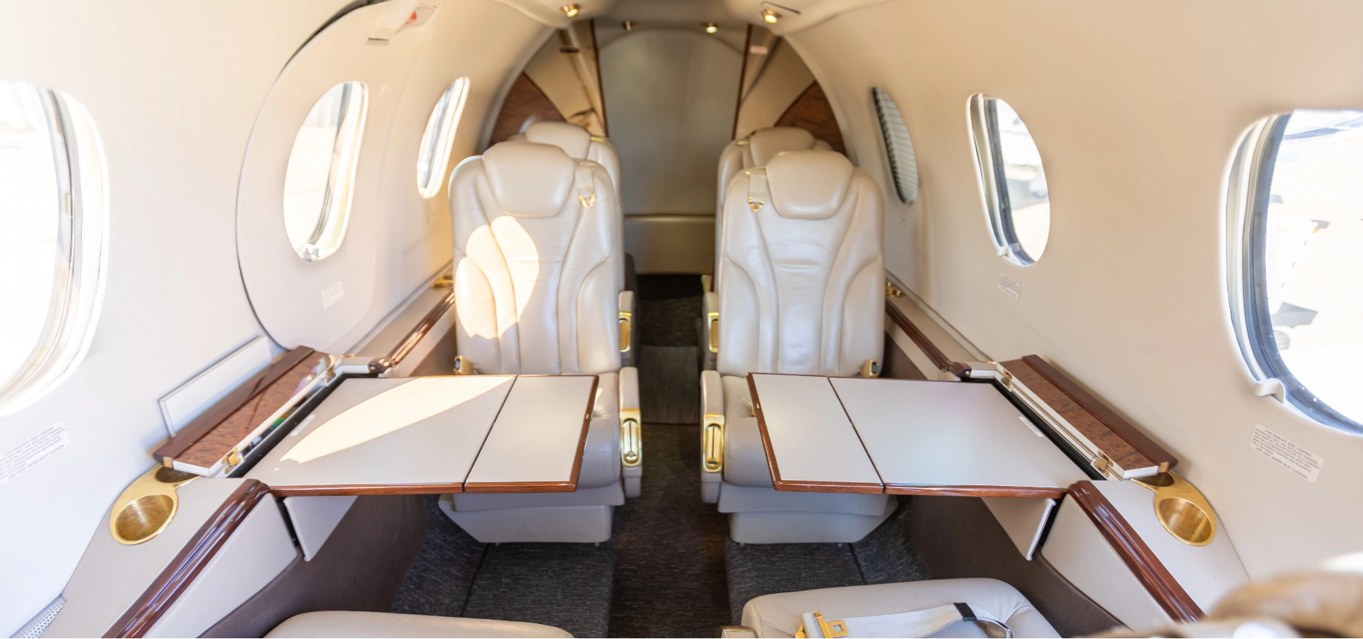 The interior of a private jet highlights luxurious aircraft acquisition, featuring four beige leather seats and fold-out tables in a spacious cabin with elegant oval windows.