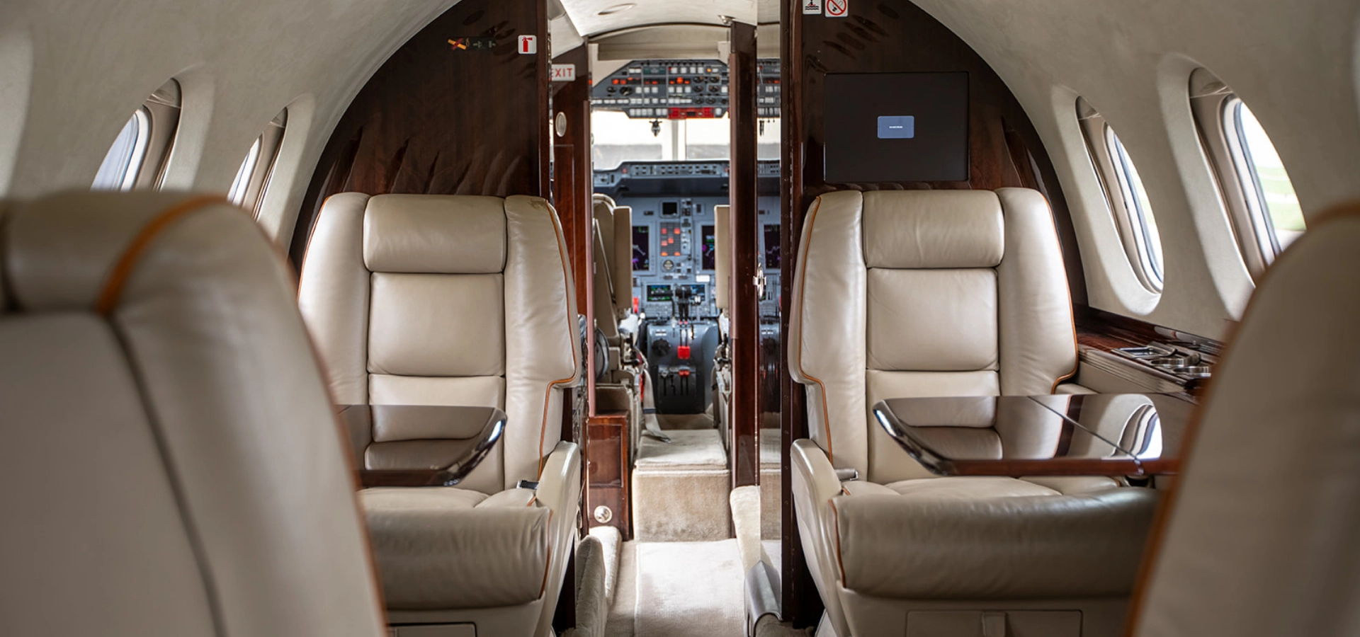 Interior view of a private jet with four beige leather seats, wooden accents, and a view towards the cockpit—perfect for those interested in luxury jet sales.
