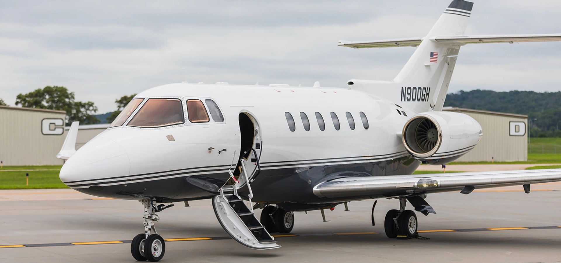 A luxury jet with an open entry door sits majestically on the airport tarmac, where hangars and greenery provide a serene backdrop—an enticing view for those exploring private jets for sale.