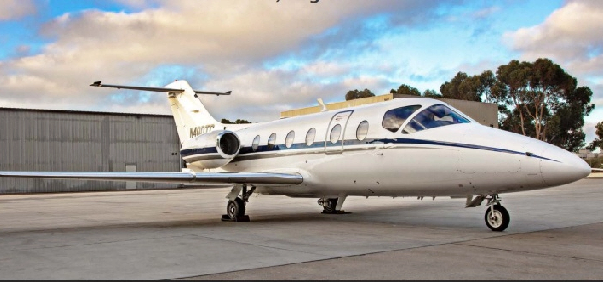A luxury jet, available for acquisition, is parked on the tarmac next to a hangar under a partly cloudy sky.