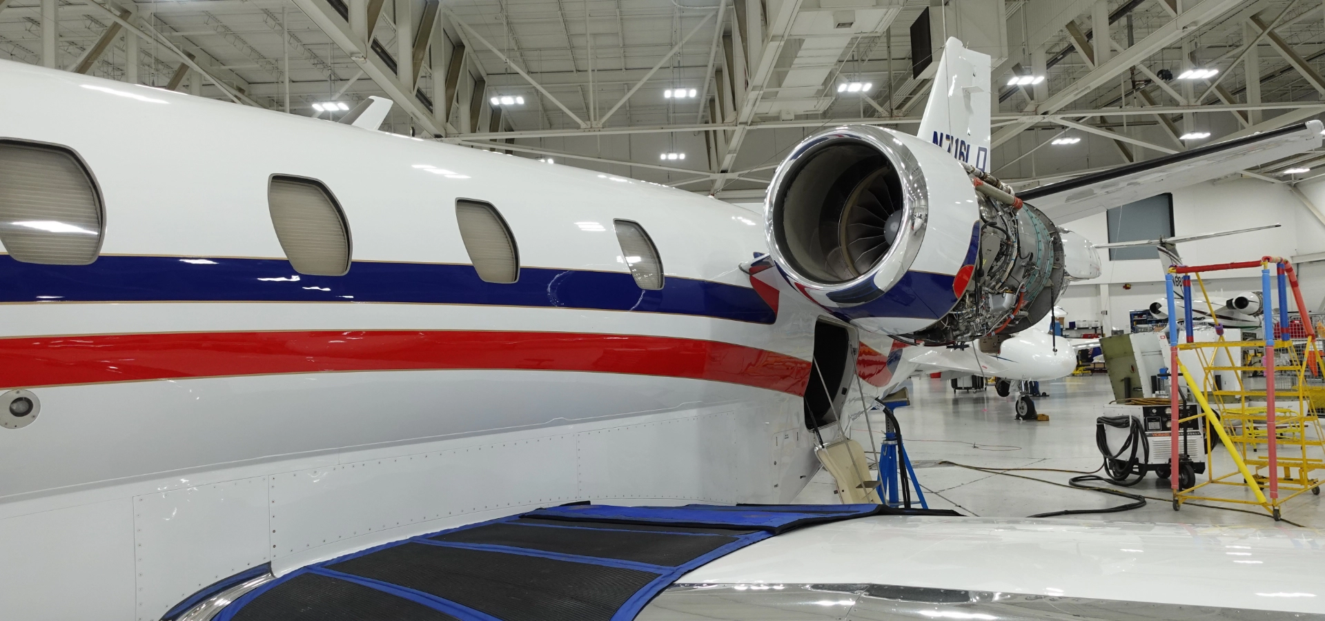 A white jet with red and blue stripes, available for luxury jet sales, is undergoing maintenance in a hangar. The engine cover is open, revealing the inner components.
