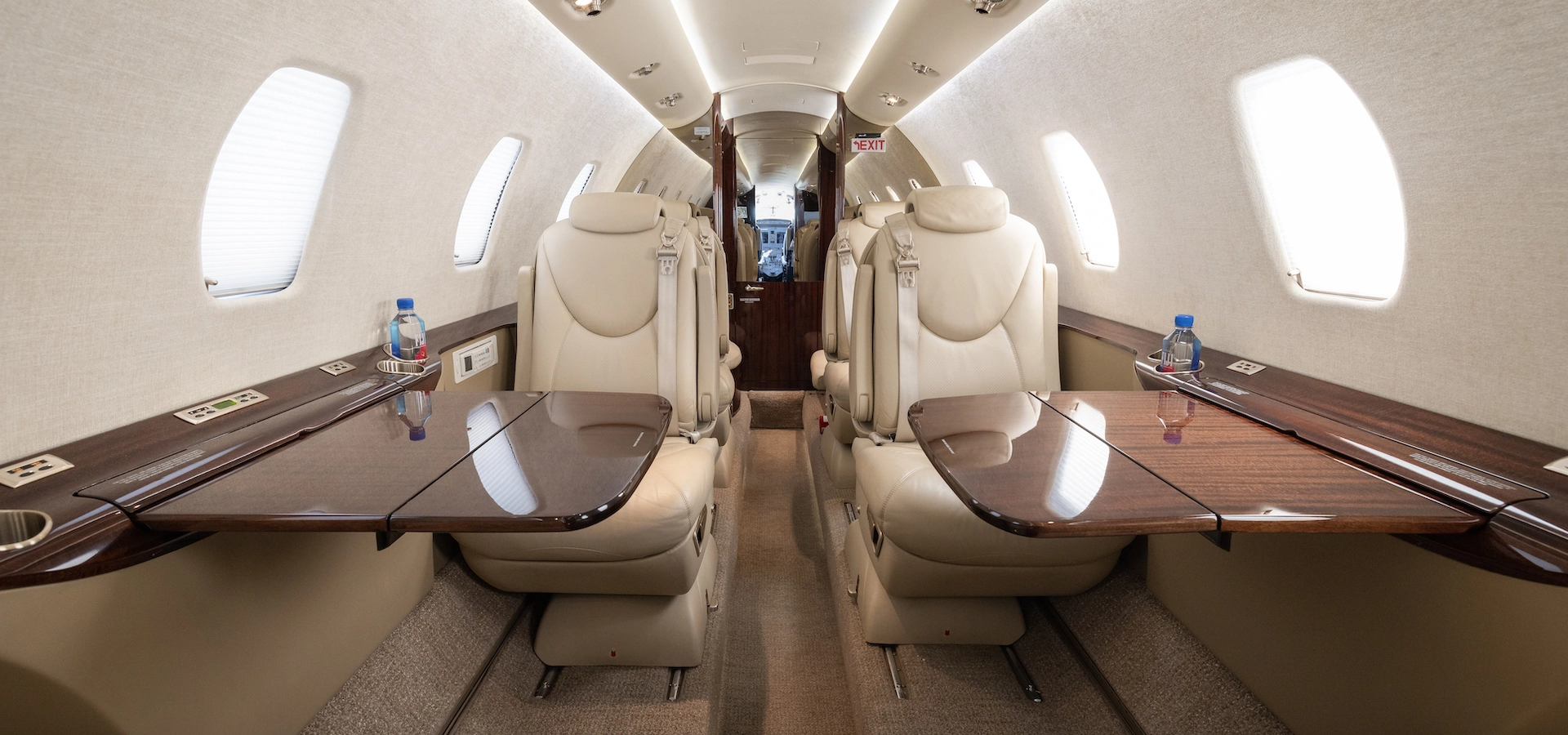 Experience the opulent interior of a corporate jet with beige leather seats, retractable wooden tables, and elegant oval windows.
