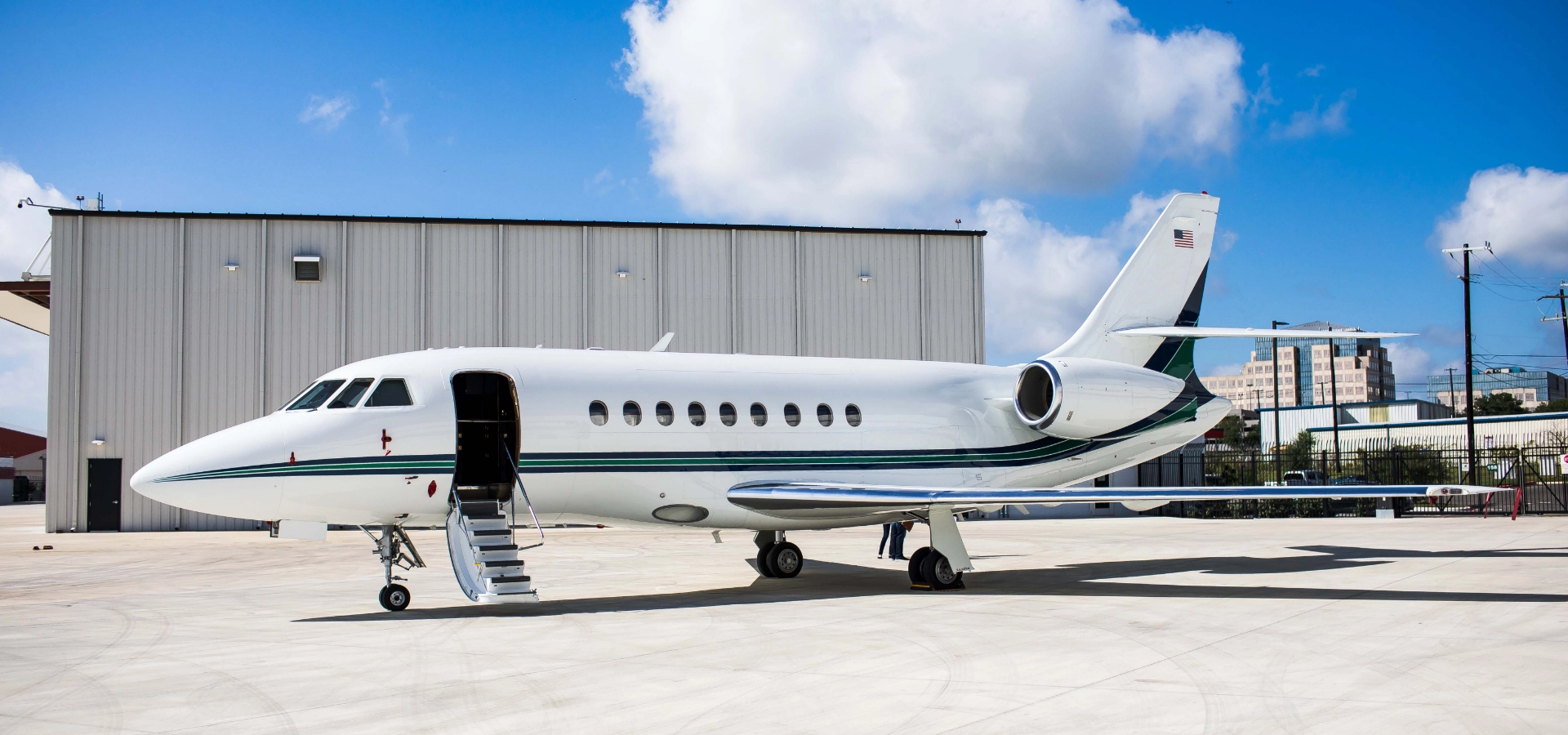 A white private jet, representative of the finest business jets for sale, is parked on an airport tarmac with a stairway extended. A large hangar looms under a partly cloudy sky, inviting thoughts of seamless aircraft acquisition.