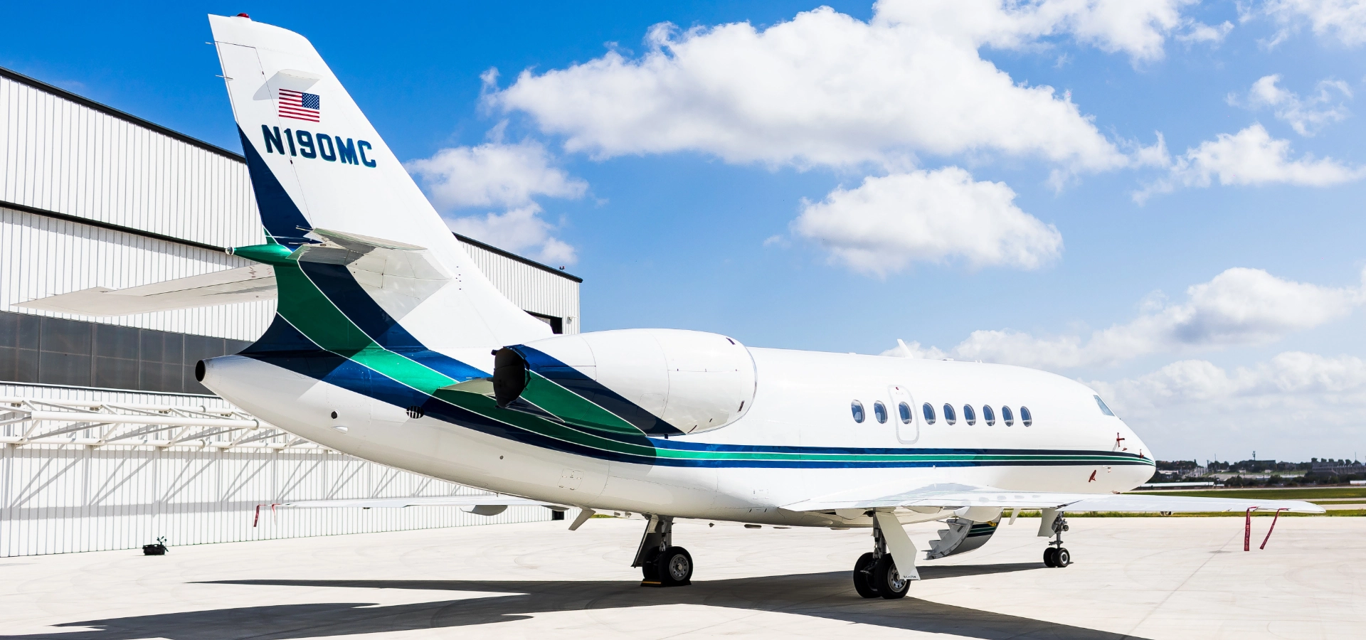 A private jet, one of the finest business jets for sale, is parked on an airport tarmac near a hangar, with a clear blue sky and clouds gracing the background.