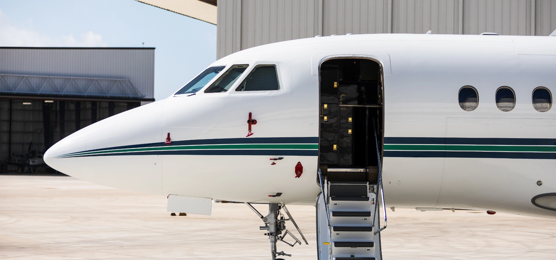 A luxury jet with an open door is parked on the tarmac near a hangar, an enticing glimpse into the world of private jets for sale.
