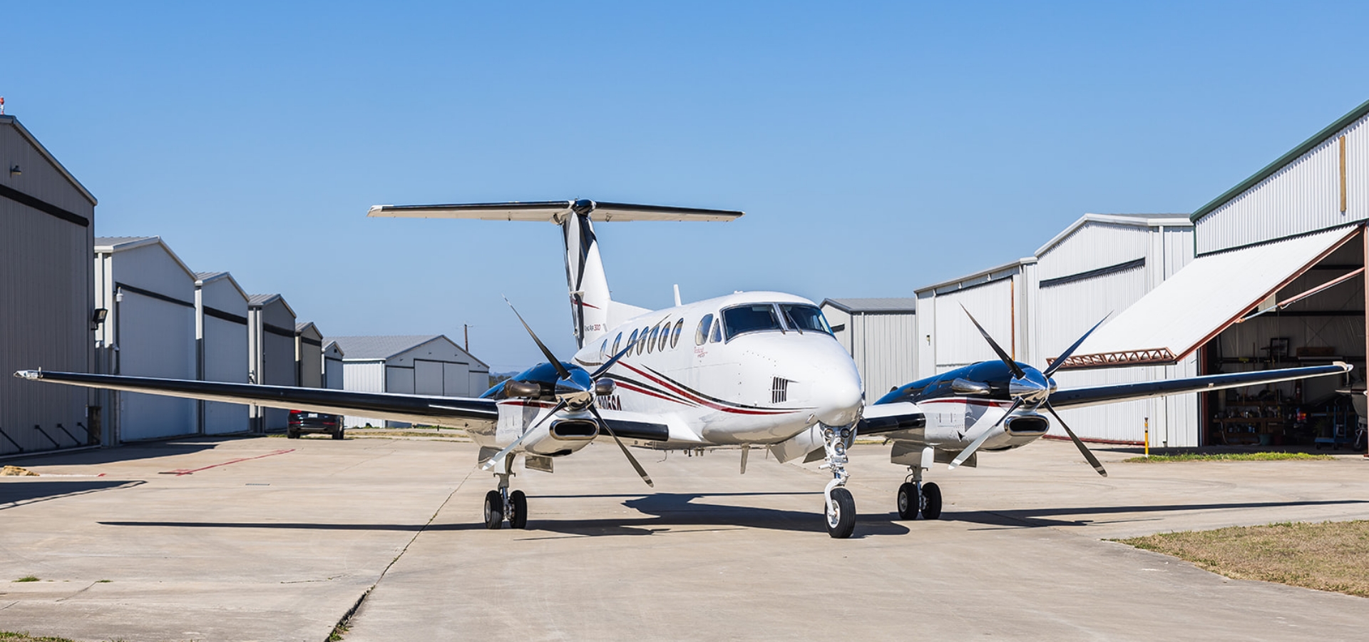 A twin-engine propeller airplane is parked on a tarmac, with hangars in the background under a clear blue sky, offering a picturesque setting for those interested in aircraft acquisition.