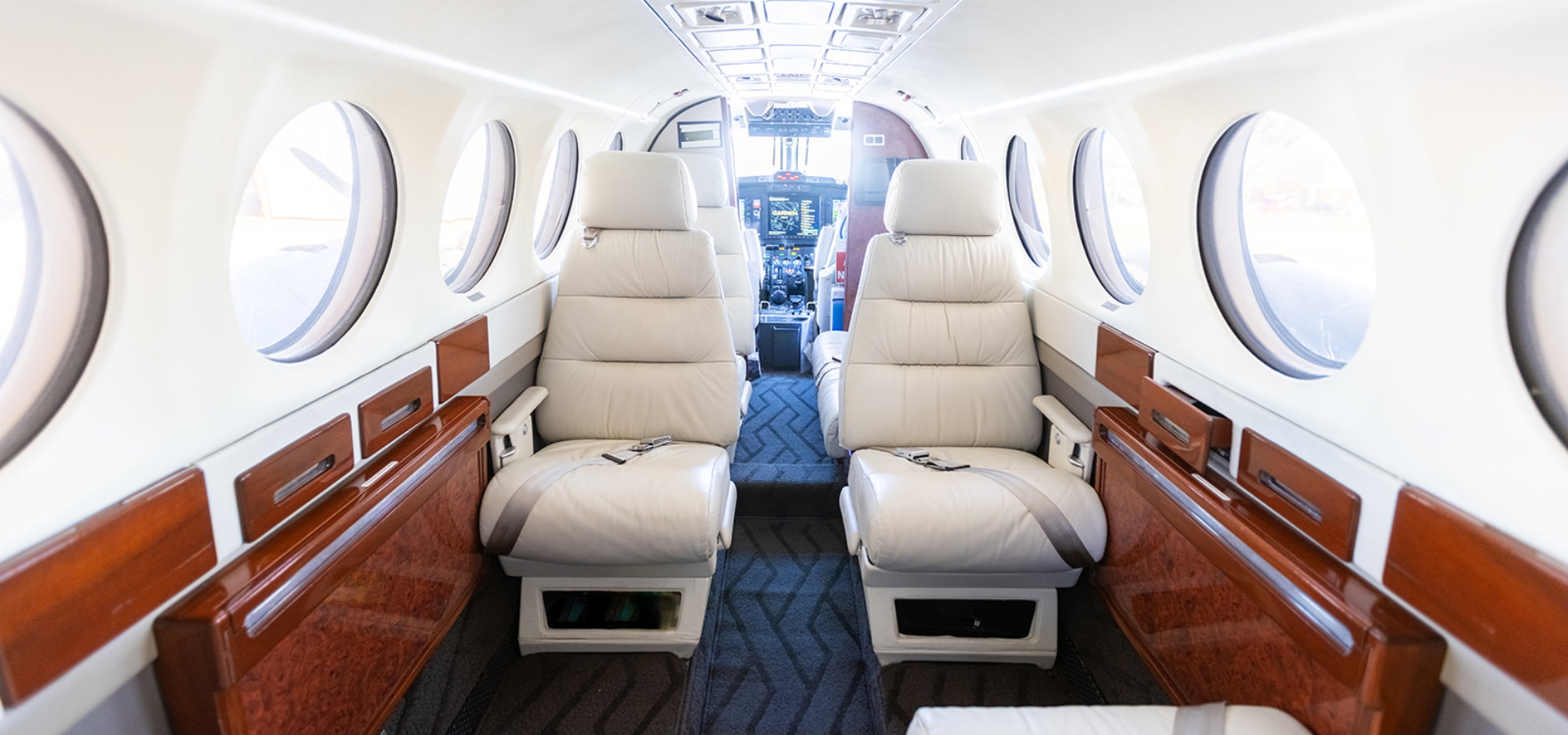 Interior of a private jet for sale, featuring two plush cream-colored seats with wood paneling, circular windows, and a view towards the cockpit.