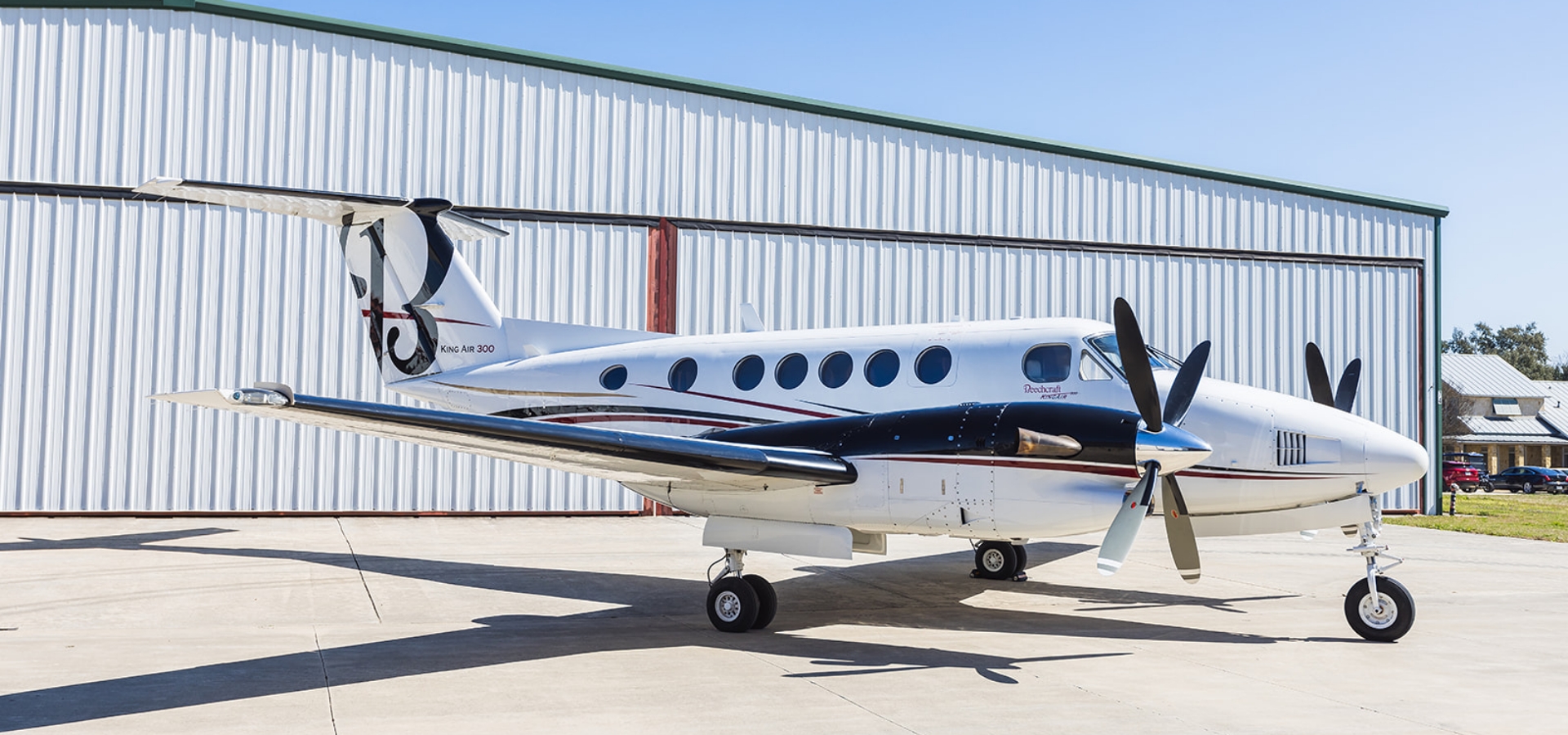 A twin-engine propeller aircraft, available for those interested in luxury jet sales, is parked on a concrete surface near a large hangar.