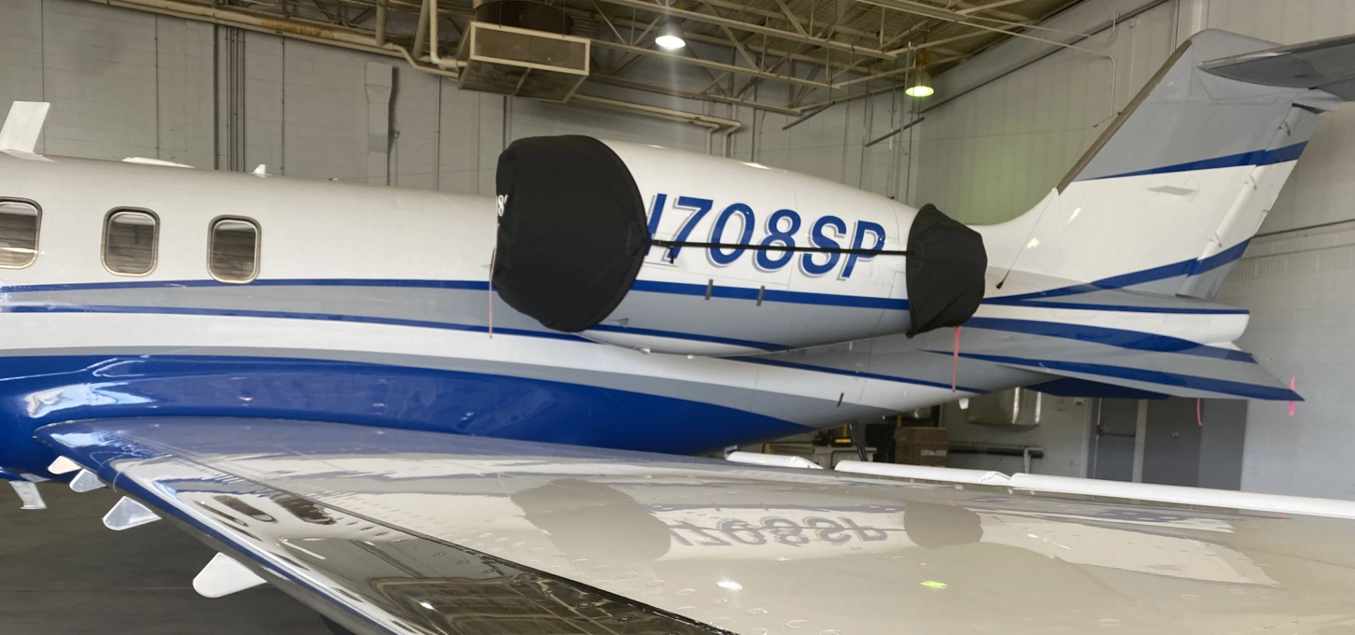 Inside the hangar, a pristine white and blue jet stands ready, showcasing its sleek partial wing and engine adorned with protective covers. Ideal for corporate jet sales, it's a testament to excellence in aircraft acquisition.