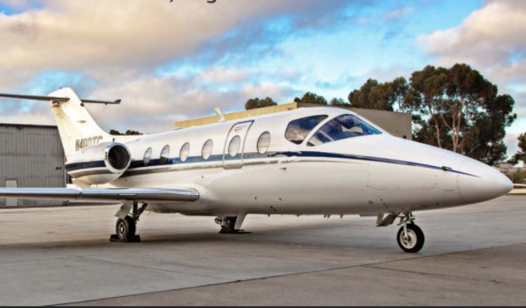 A sleek white private jet, ideal for those exploring corporate jet sales, is parked on a concrete runway. Trees and buildings provide a scenic backdrop under the partly cloudy sky, capturing the allure of exclusive air travel.