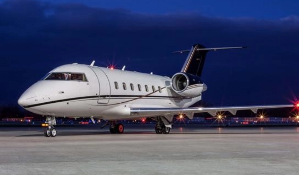 A luxury jet is parked on a runway at night with lights illuminating the tarmac in the background.
