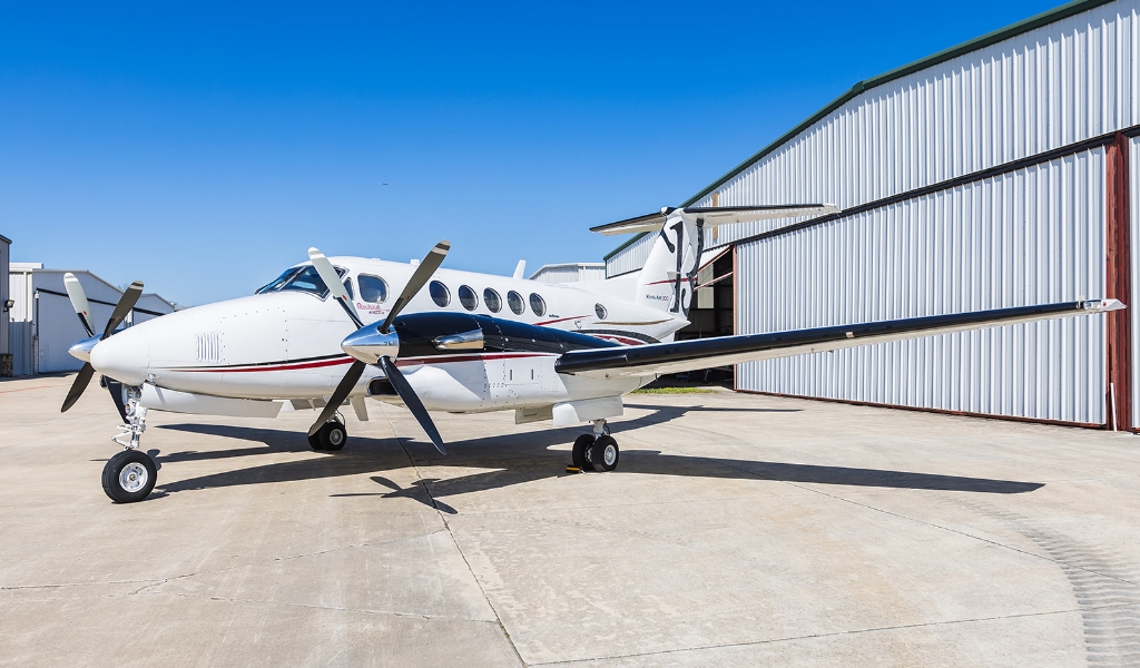 A sleek white twin-engine propeller aircraft parked on the tarmac near a hangar under a clear blue sky hints at its next adventure, potentially guided by expert aviation consulting services.
